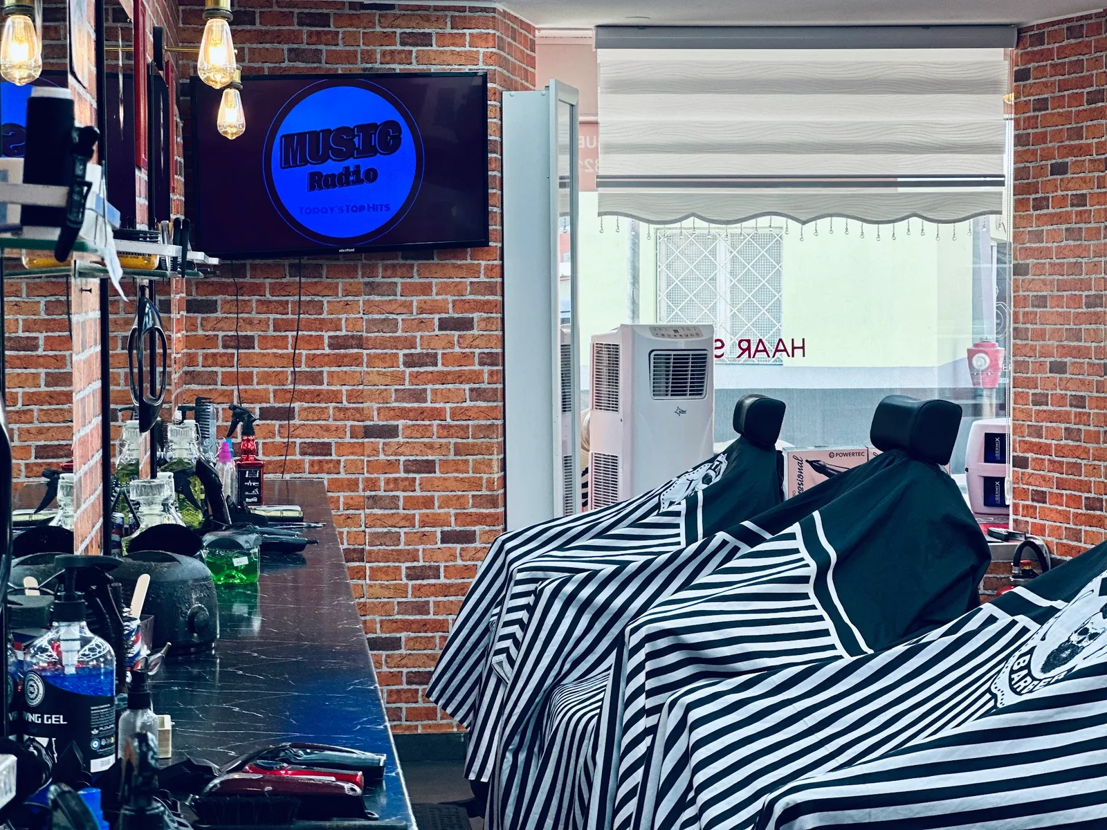 Arbeitsplatz im Salon mit Scheren, Maschinen und Pflegeprodukten auf der dunklen Ablage, daneben zwei Stühle mit gestreiften Umhängen