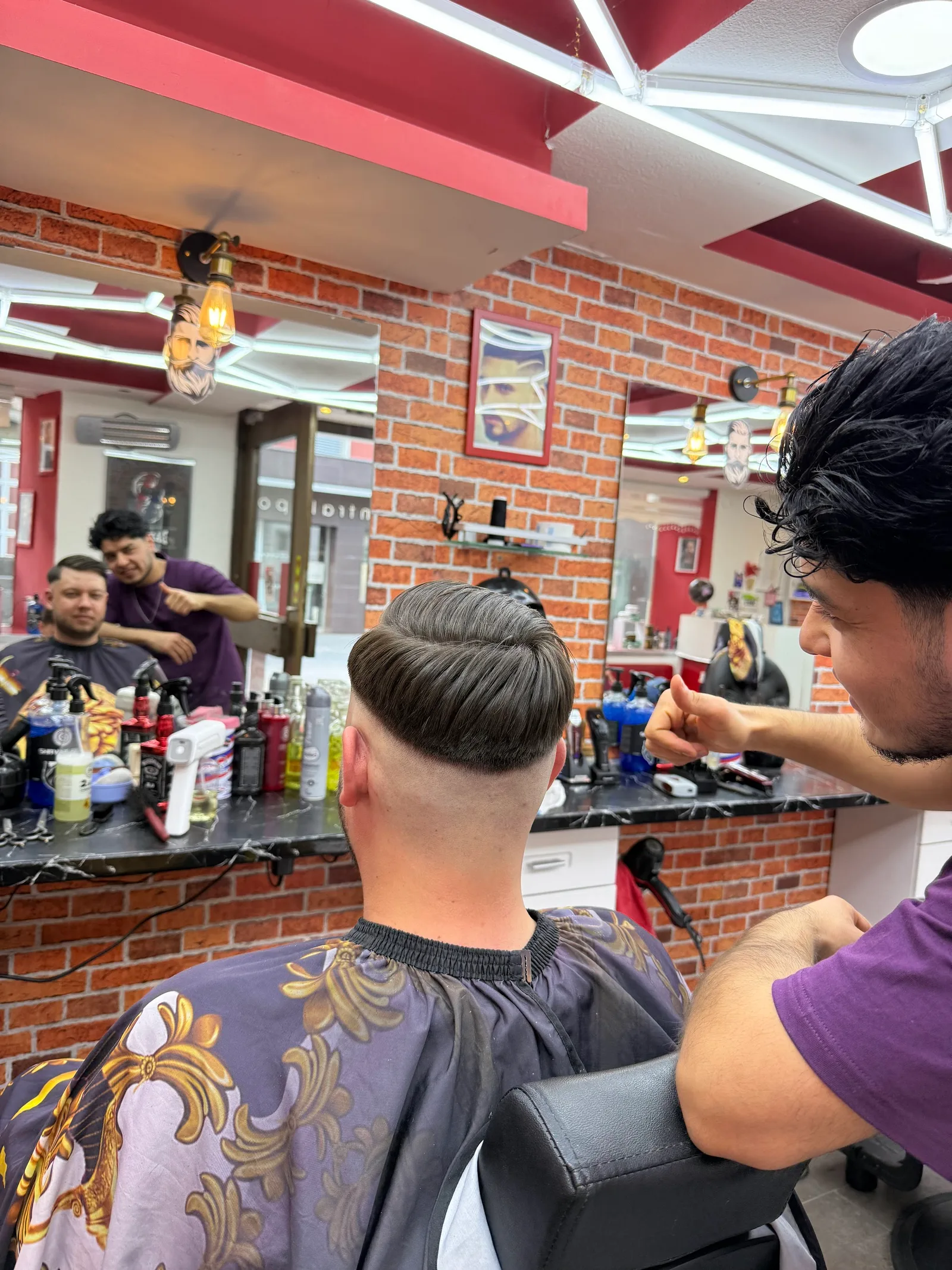 Ein Barbier zeigt dem Kunden das fertige Ergebnis, im Hintergrund der ganze Salon